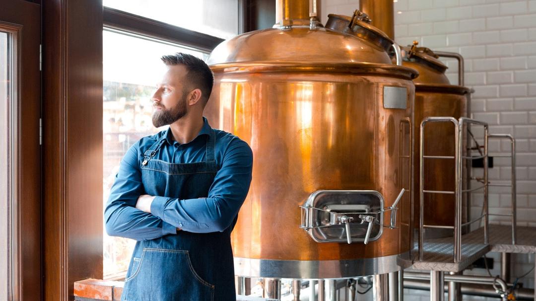 A man with a beard and crossed arms stands in front of large copper brewing tanks inside a brewery, looking out of the window. He is wearing a blue shirt and a dark apron.