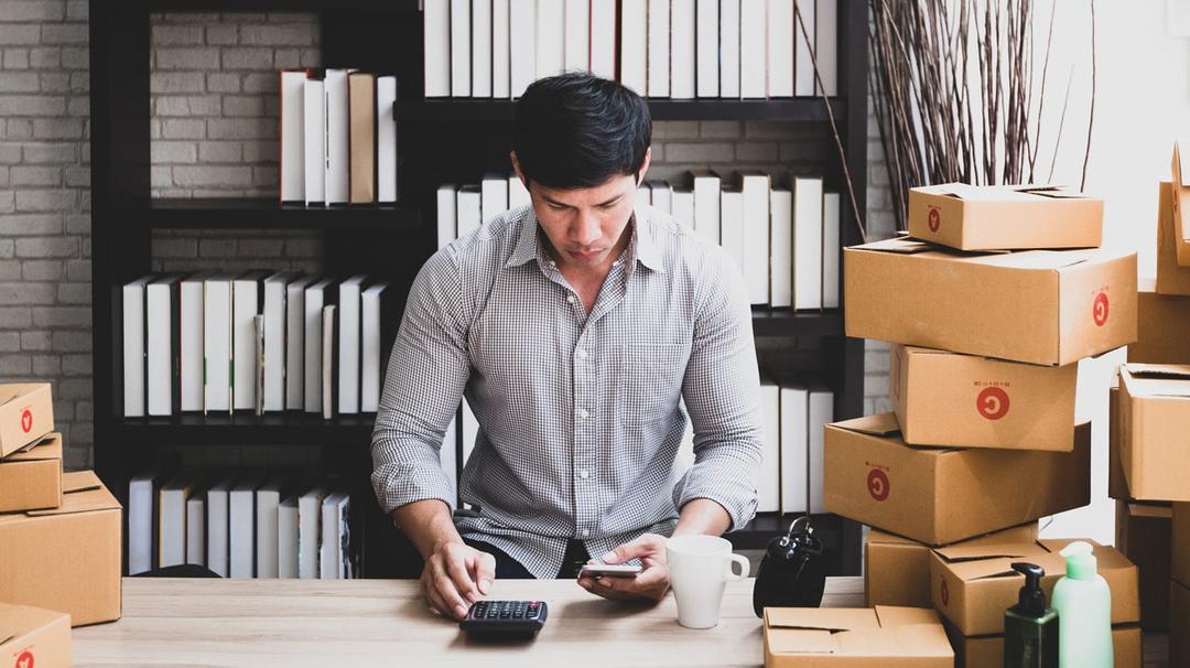 A man sitting at a desk calculating expenses with a calculator, surrounded by stacked boxes and office supplies, in a home office setting.