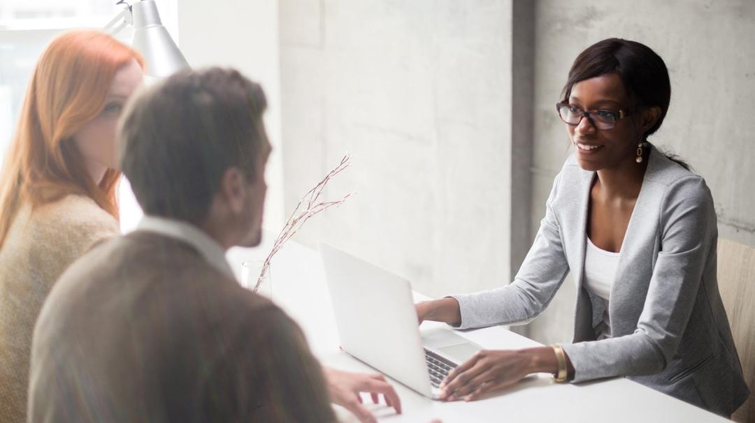 Three people are in a meeting. A woman in glasses and a gray blazer is speaking, while a man and another woman listen. A laptop is open on the table in front of them.