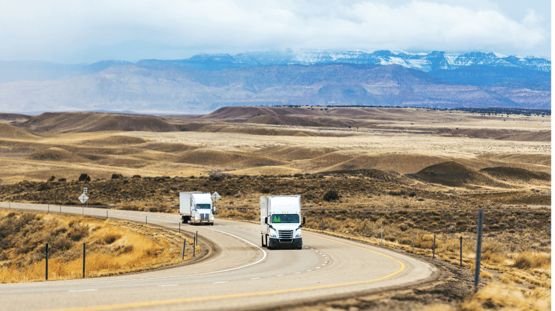 Two trucks driving on a curvy road through a desert landscape with distant snow-capped mountains under a cloudy sky.