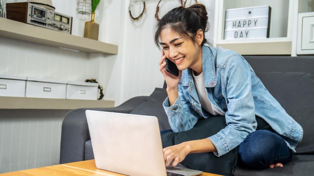 A young woman in a denim jacket smiles while talking on her phone and using a laptop in a cozy living room.