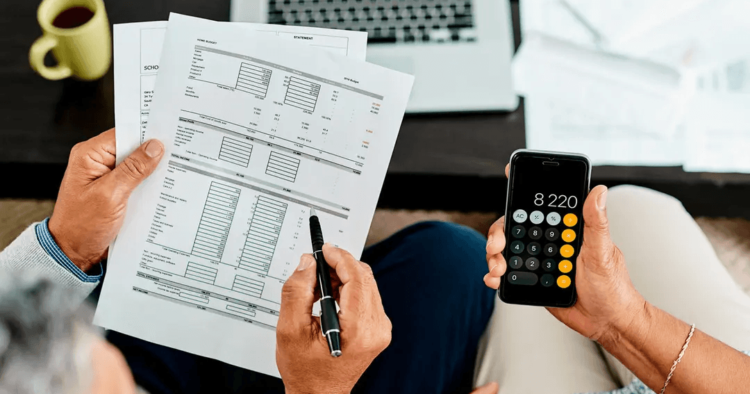 Two adults sitting, reviewing financial documents and using a calculator on a smartphone, with a laptop and papers on the table.