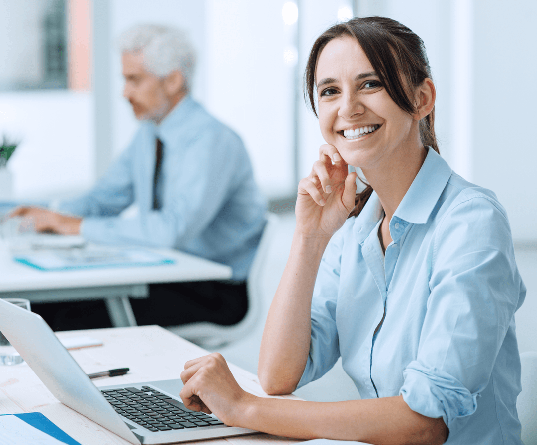 A smiling woman in a blue shirt sits at a desk with a laptop, while an older man works in the background of a bright office.